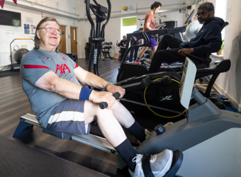 Men exercising in gym. A man on a rowing machine is in the foreground. He looks happy